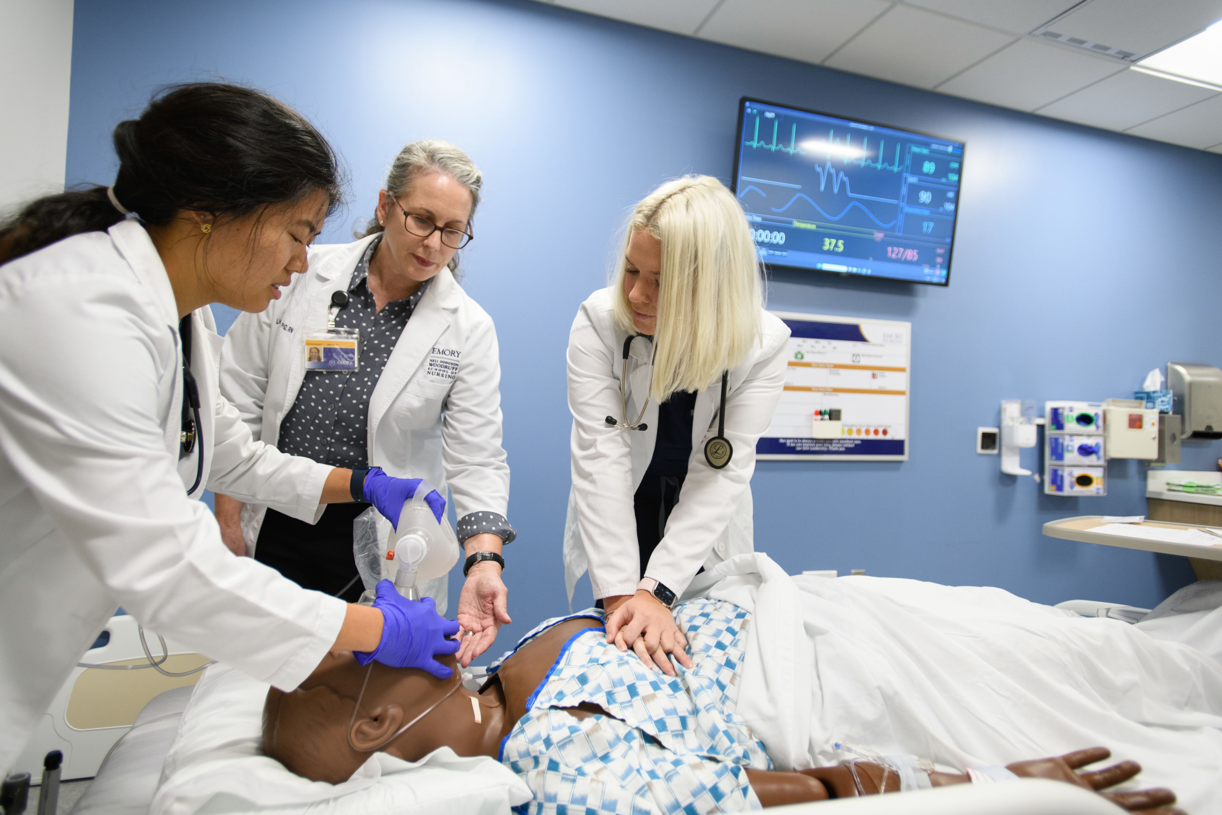 Two students performing CPR procedures on a manikin while being guided by an instructor. All three are wearing white lab coats. One student is holding a bag valve mask over the manikin's face while the other has hands held to begin chest compressions.