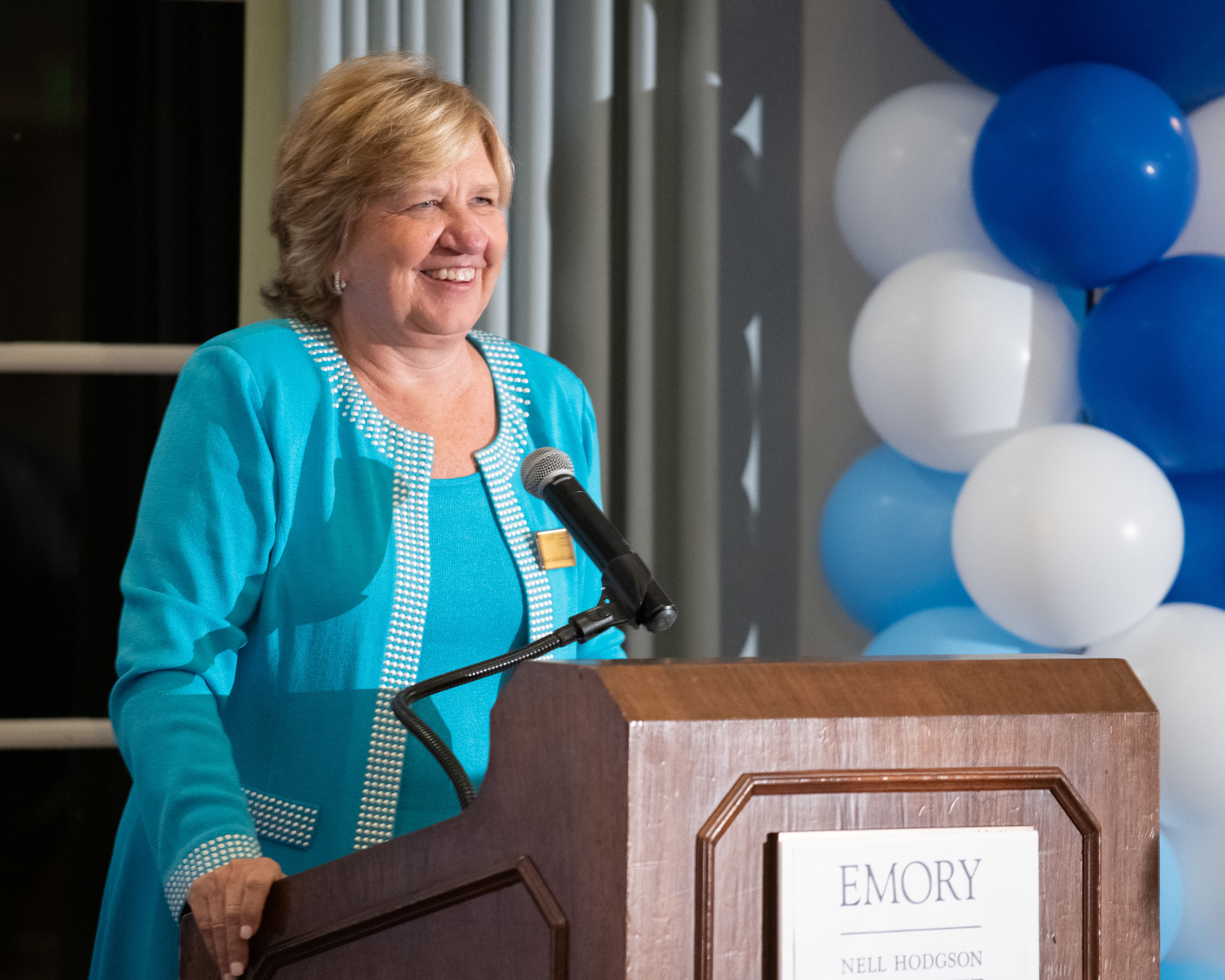 Dean McCauley in a turquoise jacket smiling at a lectern in front of blue and white balloons.