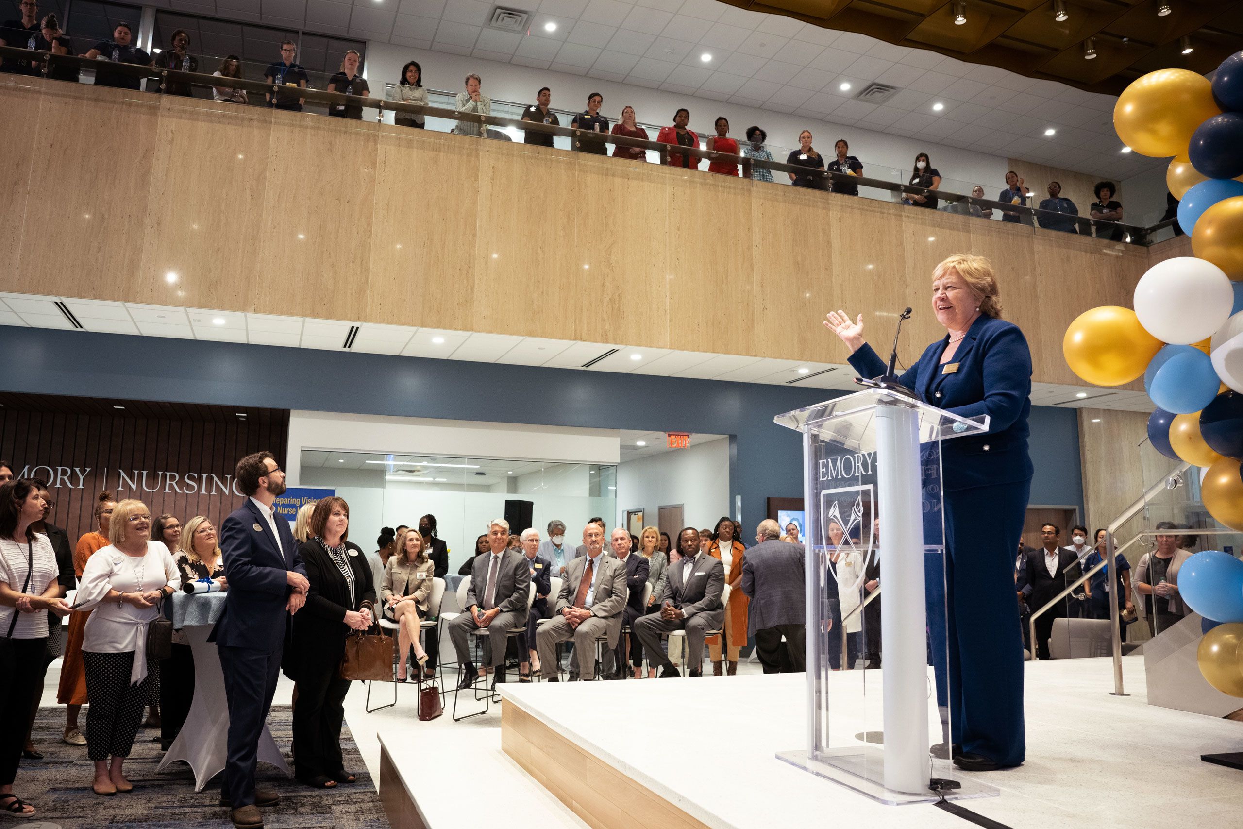 Dean McCauley speaking at a lectern in the lobby in front of a large crowd at the opening of the Emory Nursing Learning Center. There are gold, white and blue balloons throughout and the crowd is listening from two levels of the lobby.