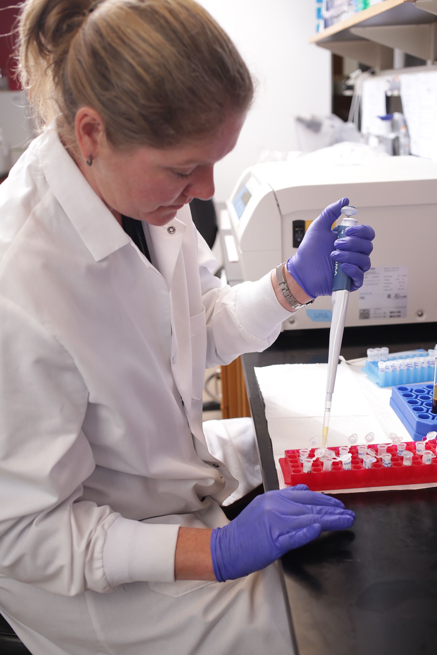 A woman in a white lab coat and purple protective gloves uses a pipette in a lab.