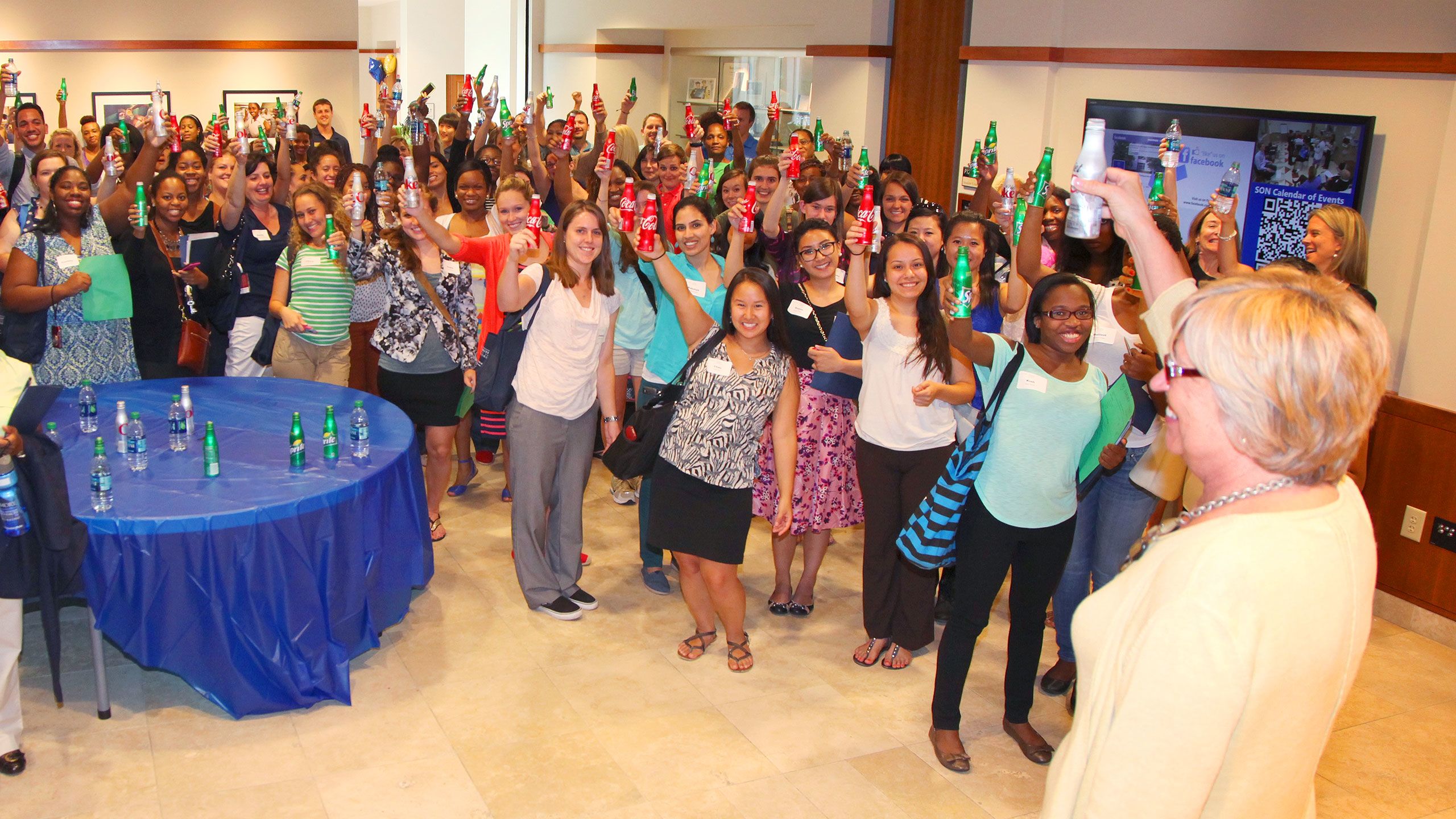 The dean stands in front of a large crowd of students and everyone is holding a bottle of Coca-Cola or Sprite up in a group toast.