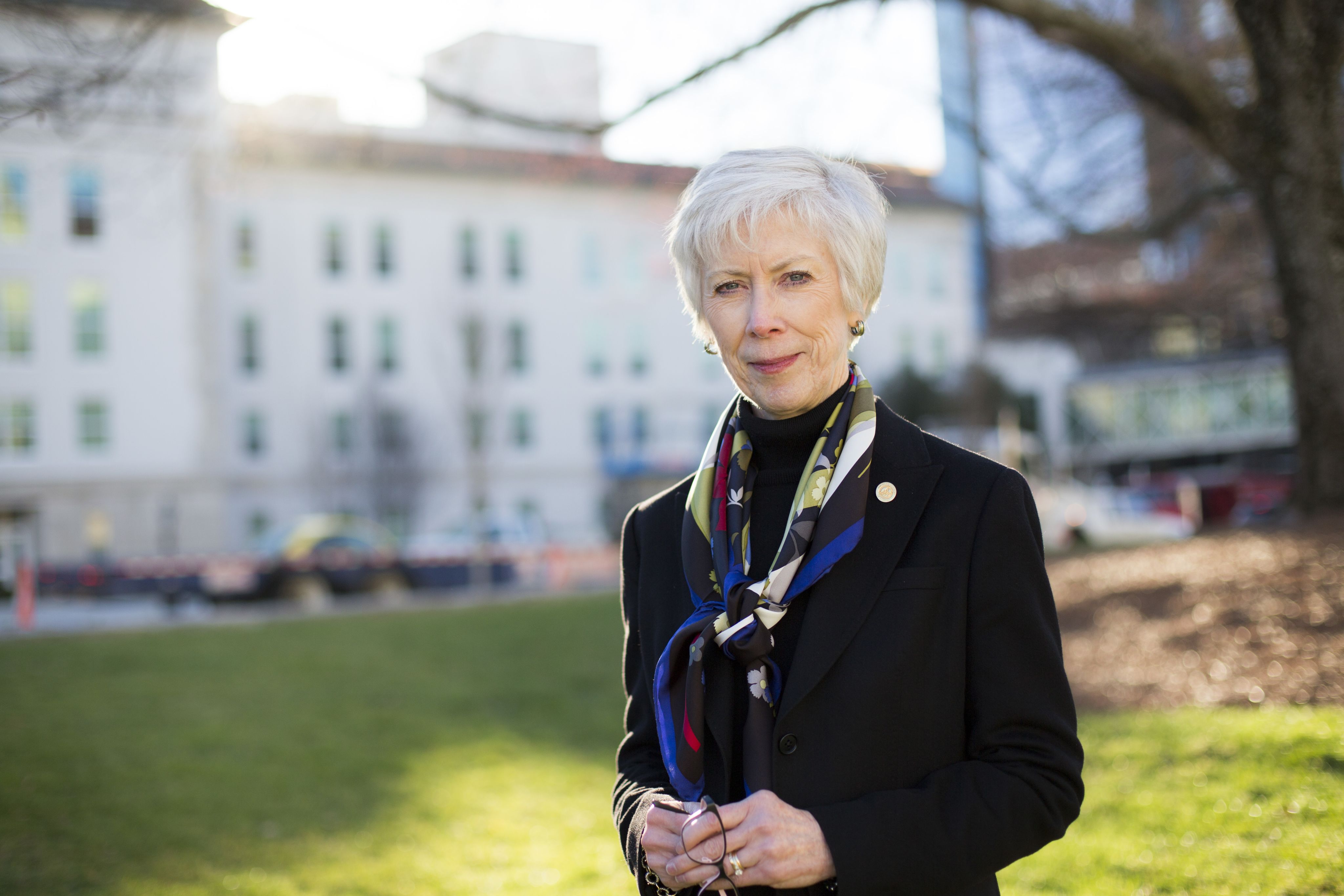 Portrait of Sharon Pappas. She has short white hair and is holding a pair of glasses. She is wearing a black blazer, a colorful scarf, and an AAN pin.