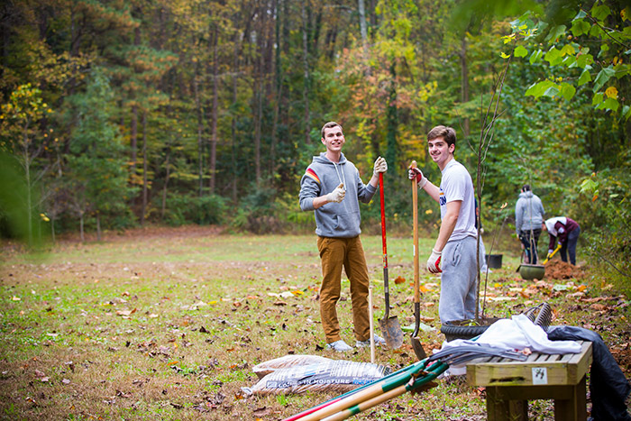 Two male students pose with large shovels as they help prune the community garden.