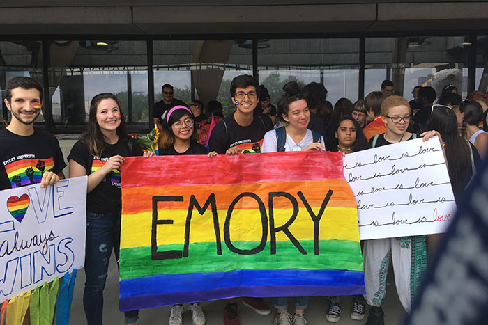 A group of Emory students holds up a large, homemade sign with the word "Emory" on it and a rainbow background.