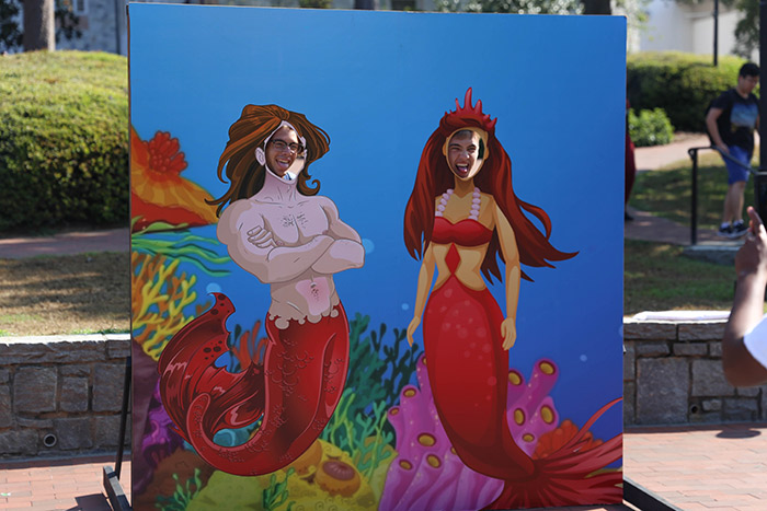 Two students pose with their faces showing through a large illustration of two mermaids.