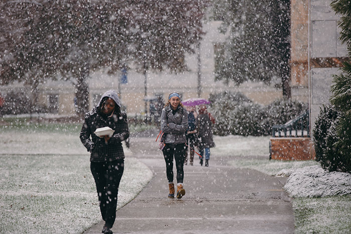 One student carries her lunch and another smiles as their walk across campus includes snowfall.