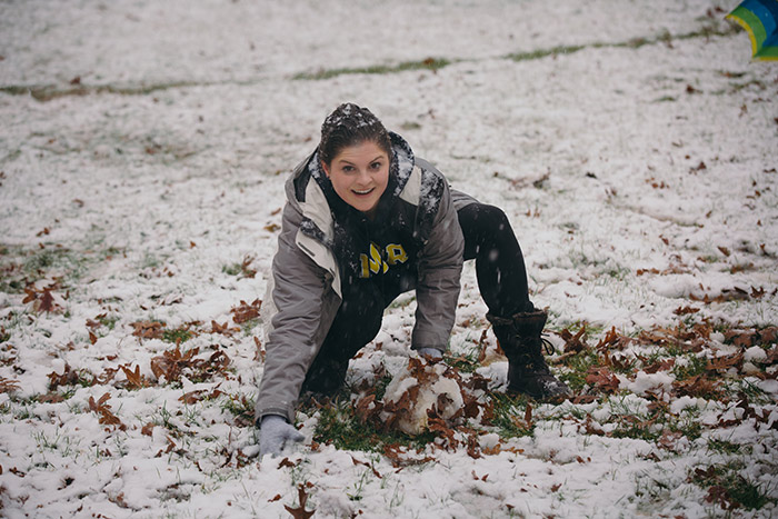 A student kneels on the ground and grins as she starts to roll snow into a ball.