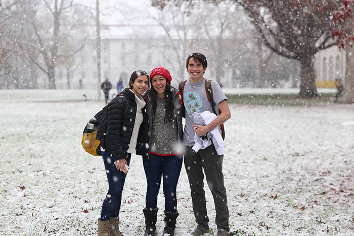 Three students gather together and smile as the snow falls on campus.