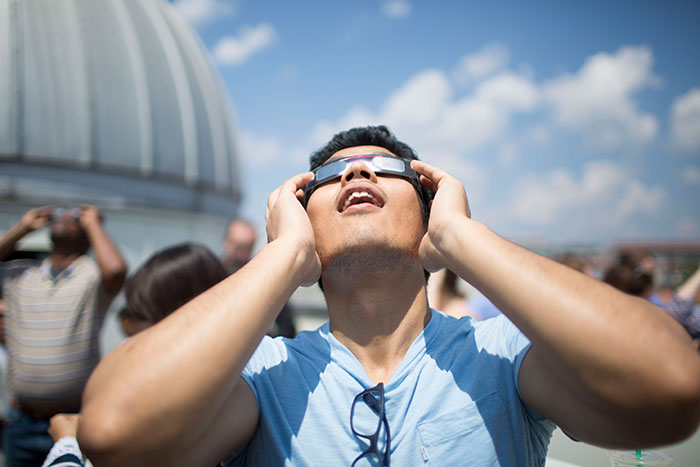 A student looks above through eclipse glasses at the sky.