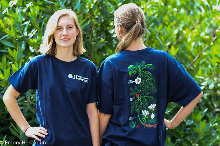 Two female students model Emory blue t-shirts with a plant illustration running across the back of the shirt