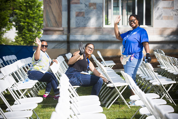Three Emory employees wave at the photographer as they help set up chairs in the quad