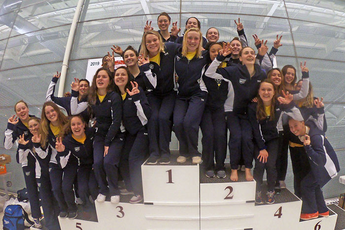 The Emory women's Swimming & Diving team stands on a riser together, posing for a champions photo in matching Emory Athletics jackets