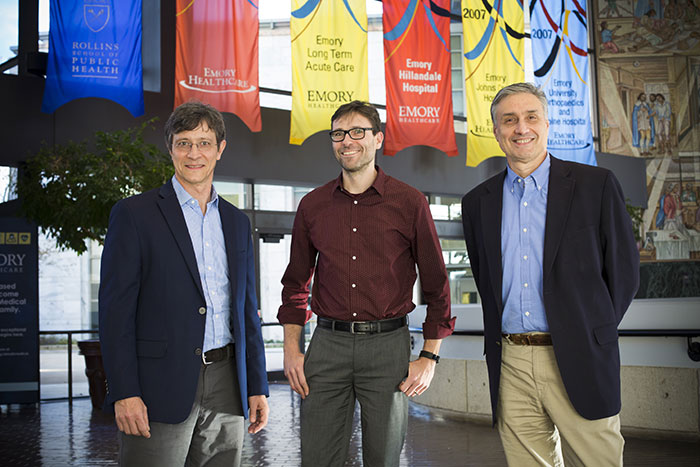 Ken Moberg, Jaap de Roode and Guido Silvestri pose together in front of several Emory banners