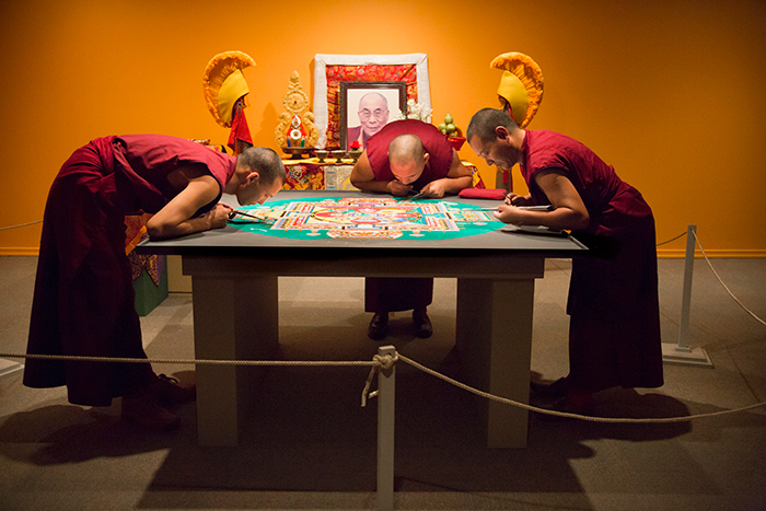 Three monks working on mandala with dalai lama in background