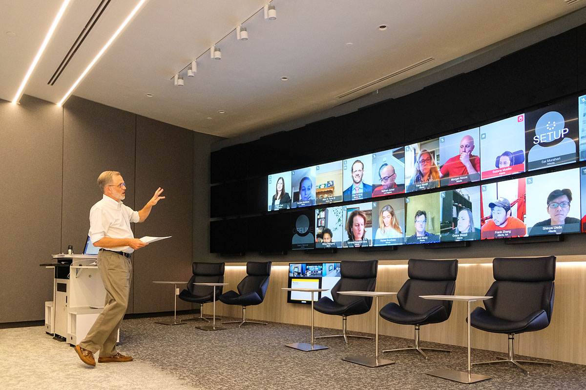 A professor teaches a class to students who are attending virtually on a large screen in a classroom