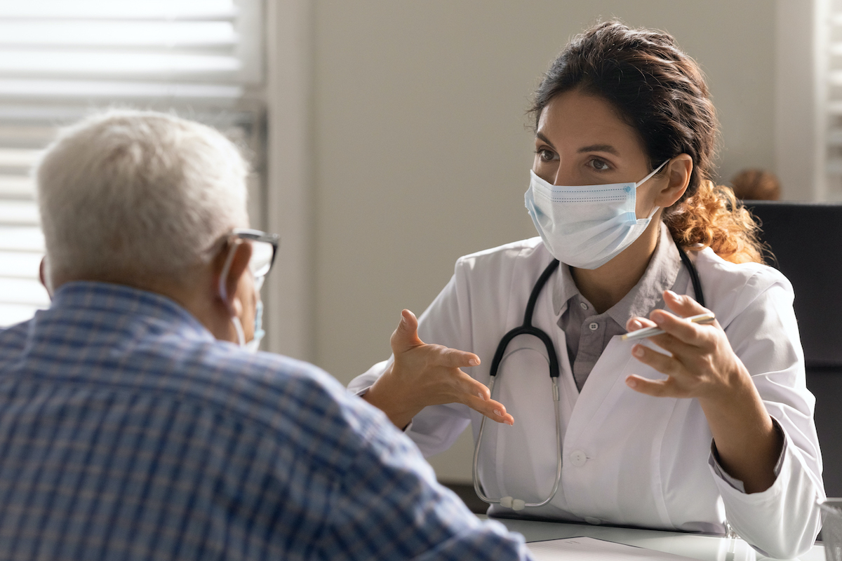 A doctor wearing a mask, stethoscope and lab coat speaks to a patient wearing a mask.
