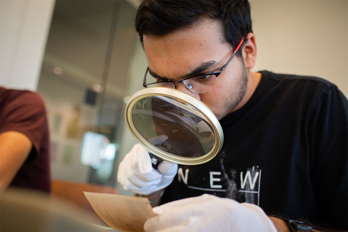 male college student holding magnifying glass to examine historic library texts