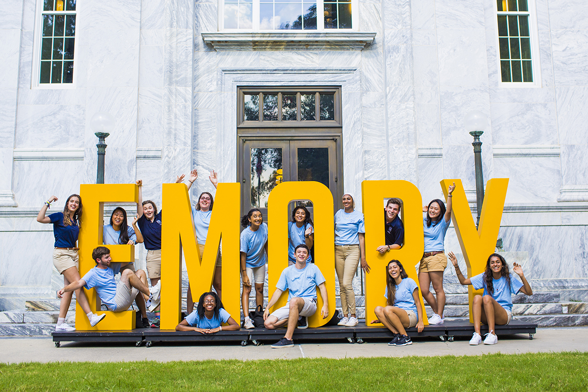 Photo of students smiling and waving while standing inside large EMORY sign