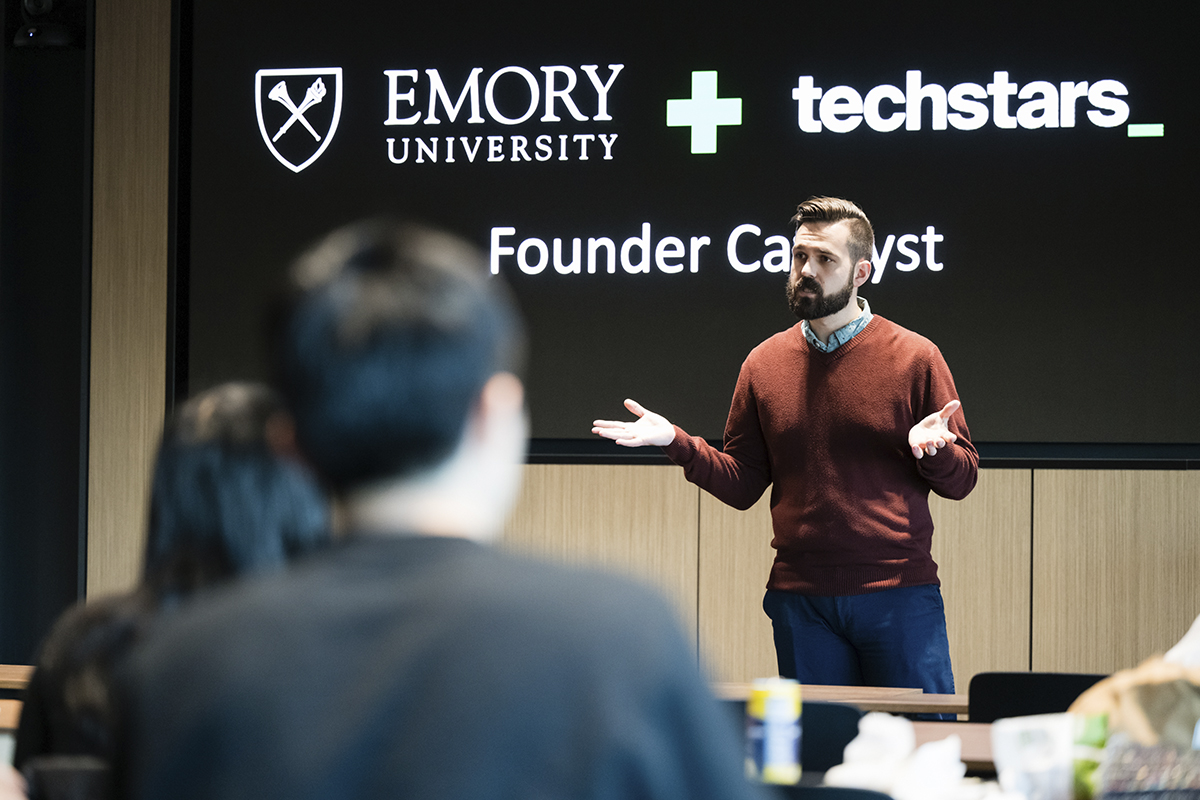 man presenting a talk in front of a board with the words emory plus techstars