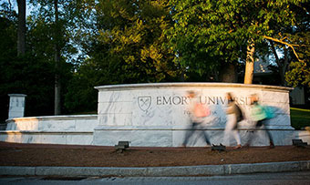 Three students walk quickly in front of the Emory sign