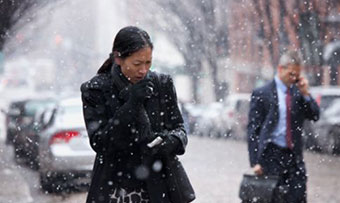 A woman wrapped in a winter coat coughs on a busy city sidewalk