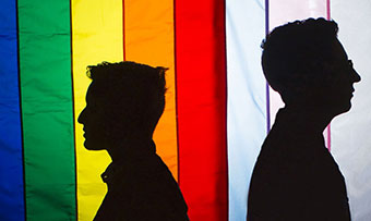 Two silhouettes stand back to back in front of a rainbow flag and a pink, blue and white-striped flag