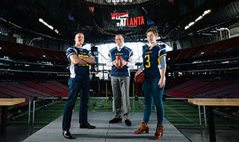Three Emory faculty members pose in Mercedes-Benz stadium, wearing Emory football jerseys